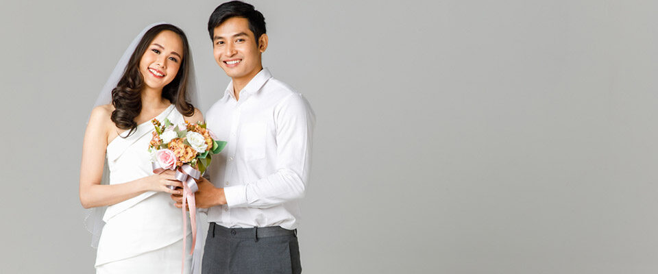 Young attractive Asian couple, man wearing white shirt, woman wearing white dress with wedding veil holding bouquet of flowers together. Concept for pre wedding photography.